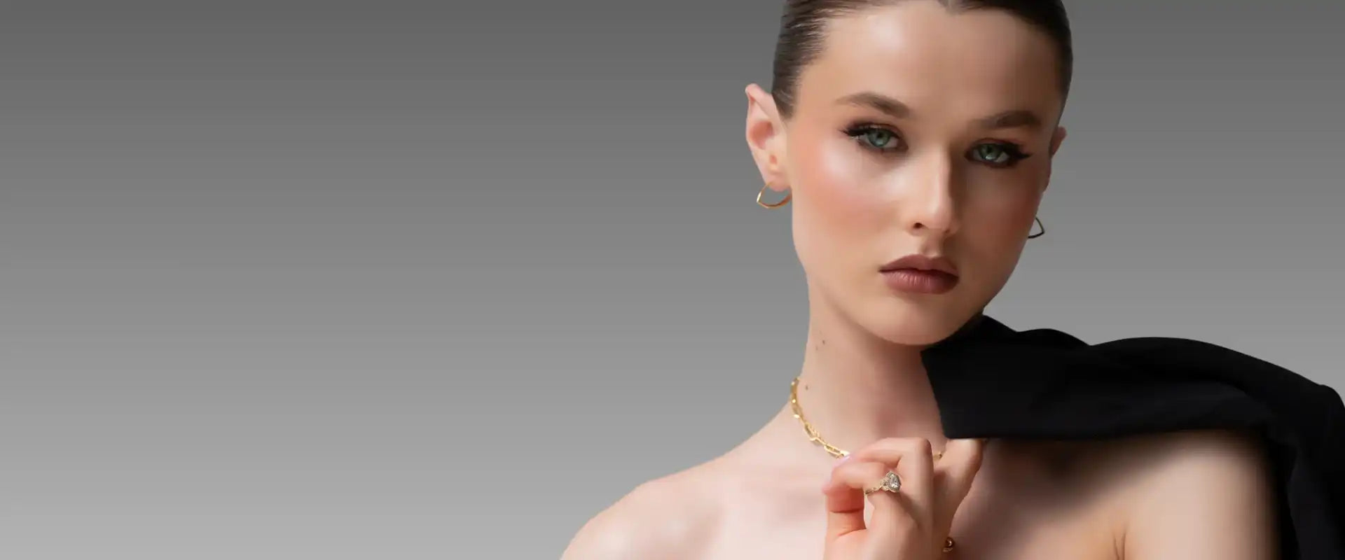 Woman with green eyes wearing gold hoop earrings, chain necklace, and diamond engagement ring, holding black jacket